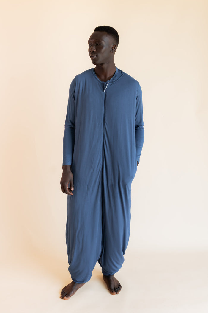 A man wearing a dusty blue sleep sack with a front zipper, standing against a solid beige background.