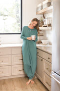 A woman wearing a sage green sleep sack stands barefoot in a bright kitchen, holding a white mug.