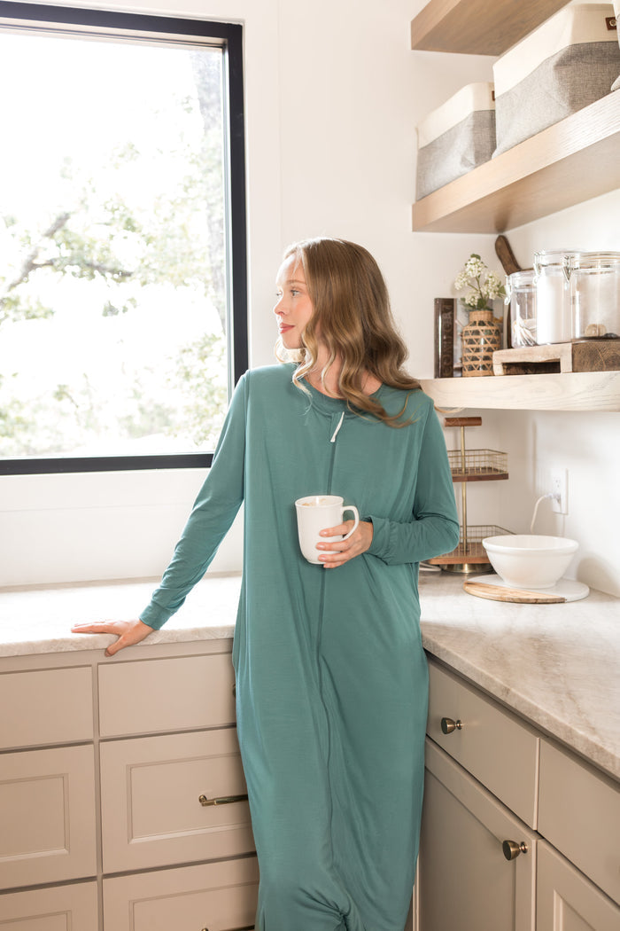 A woman wearing a sage green sleep sack in a bright kitchen, holding a white mug.