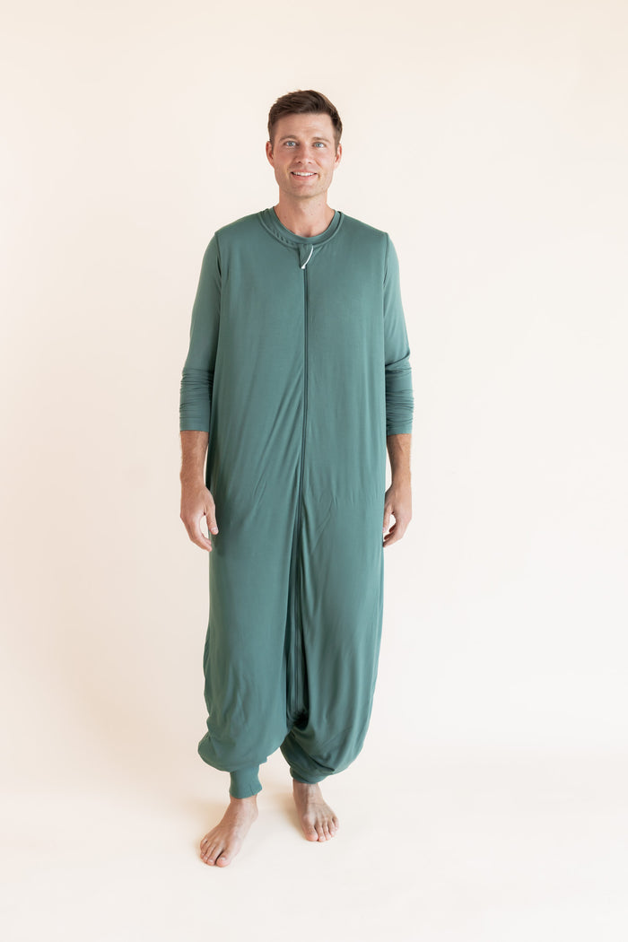 A man wearing a sage green full-body zip-up sleeper sleep sack, standing against a plain off-white studio background.