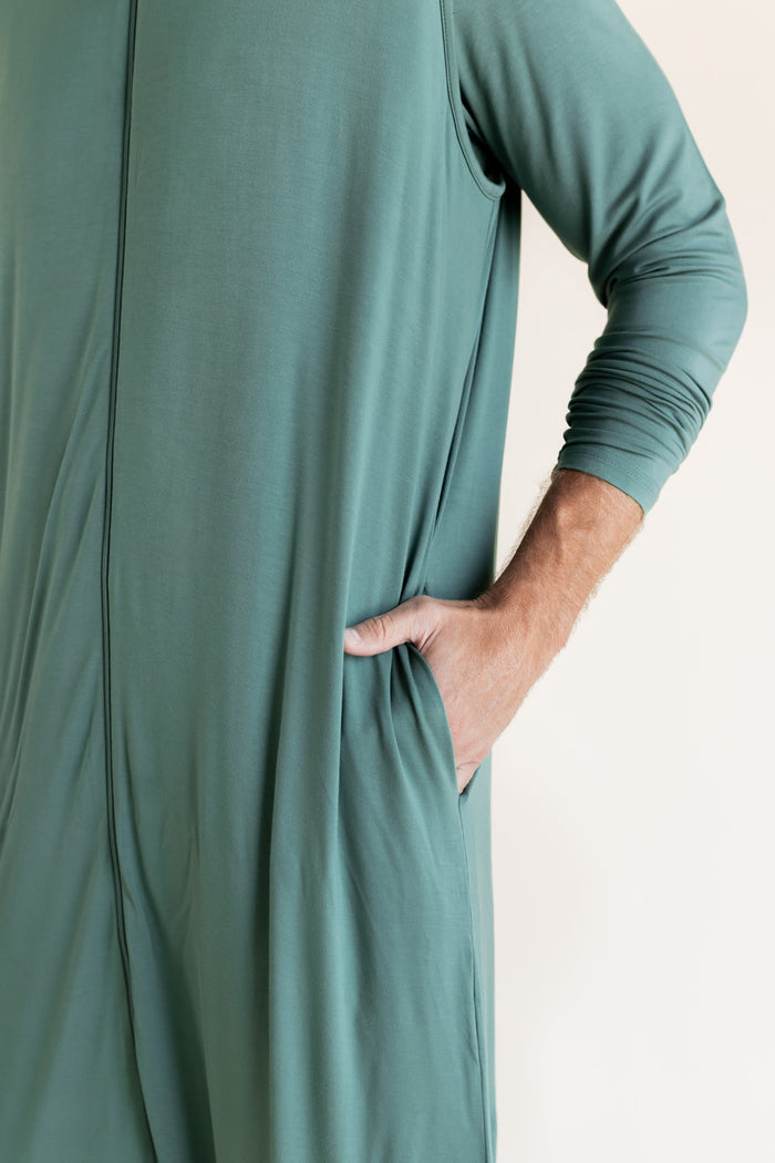 A close-up of a man wearing a sage green sleep sack, with his hand in a side pocket, against a plain off-white background.