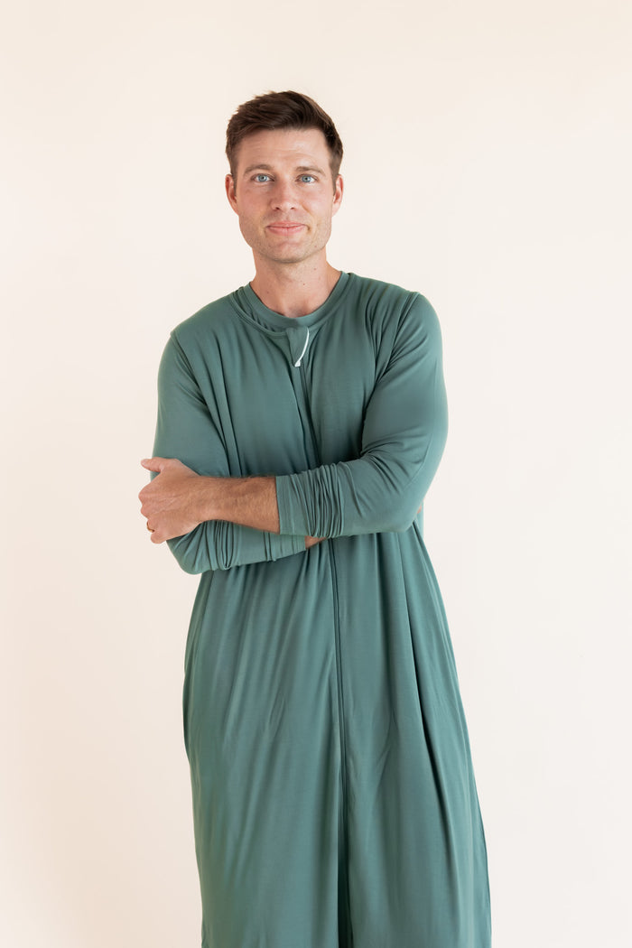 A man wearing a sage green sleep sack, standing with his arms crossed against a plain cream-colored background.