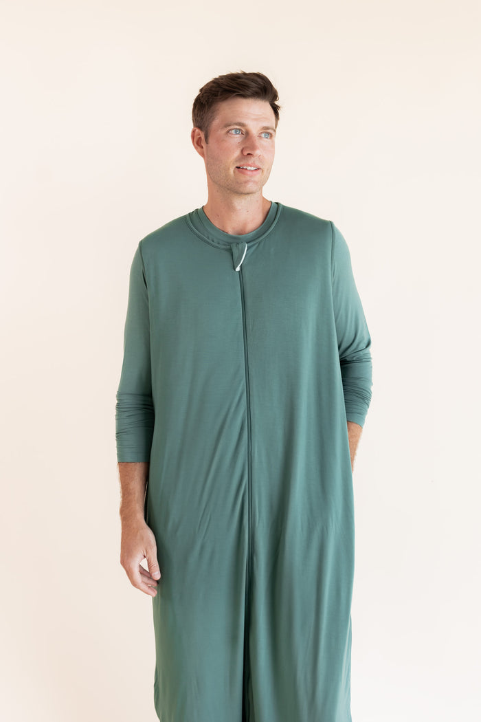 A man wearing a sage green sleep sack with a front zipper, standing against a plain cream background.