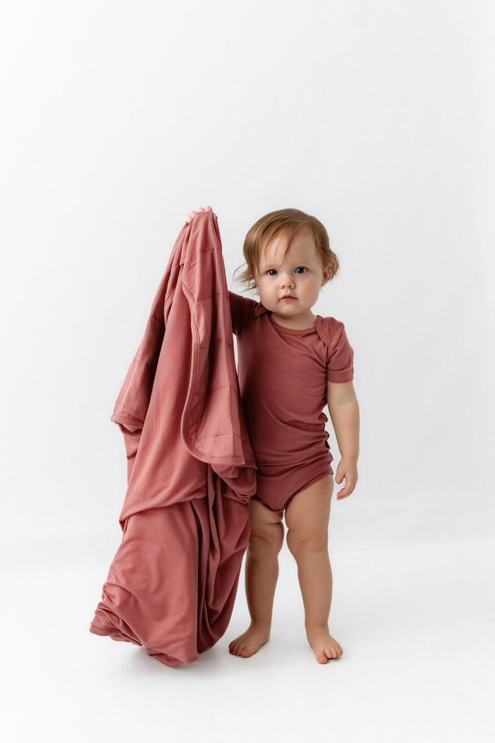Baby holding Baby Blanket in Ember while wearing a similar color bodysuit dress on a white background