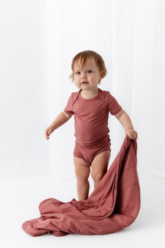 Baby holding Baby Blanket in Ember while wearing a similar color bodysuit dress on a white background