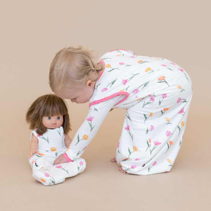 Young girl wearing the Sleep Bag Walker in Tulip 0.5 leaning over a doll sitting on the floor wearing the baby doll sleep bag in tulip