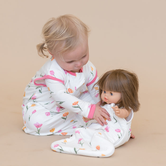 Toddler siting on the floor wearing the Baby Doll Sleep Bag in Tulip playing with a doll wearing the baby doll sleep bag in Tulip