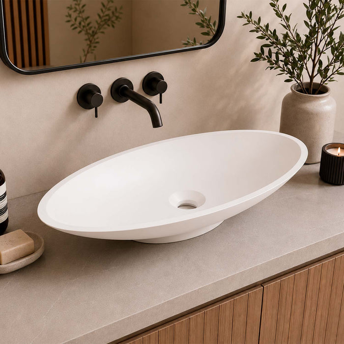 An oval white vessel bathroom sink with a matte finish, set into a gray stone countertop with a black faucet, a mirror, a potted plant, and a candle in a styled bathroom setting.