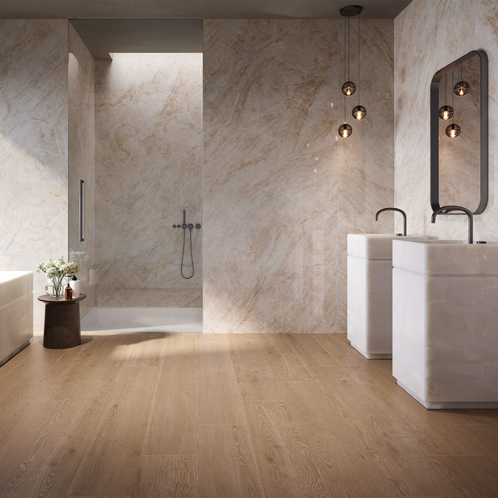 A modern bathroom interior with light wood flooring, beige marble walls, a walk-in shower, two white pedestal sinks with dark faucets, and a black-framed mirror.
