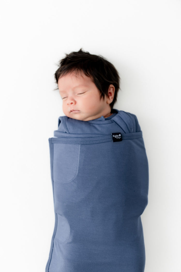 A sleeping newborn baby wrapped in a dusty blue swaddler, shown against a plain white background.