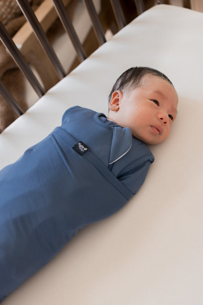 A newborn baby lying on a white mattress, swaddled in a blue sleep bag swaddler.