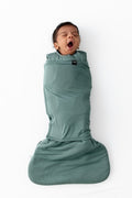 A baby wrapped in a sage green swaddler, yawning while lying on a white background.