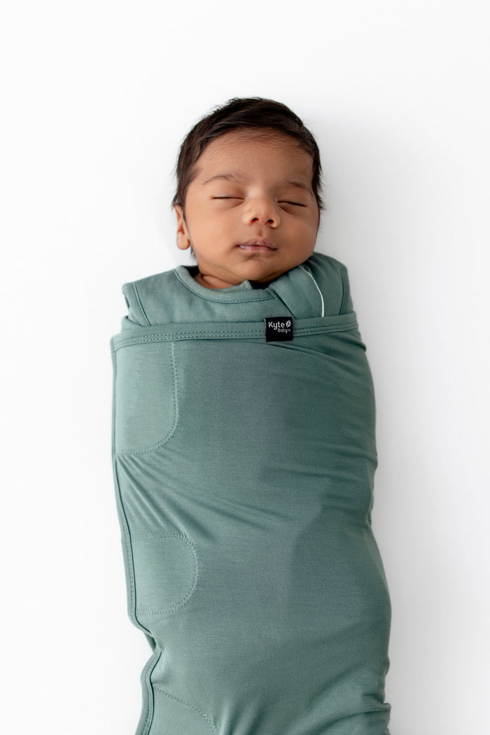 A sleeping newborn baby swaddled in a sage green sleep bag swaddler, shown from the side against a plain white background.
