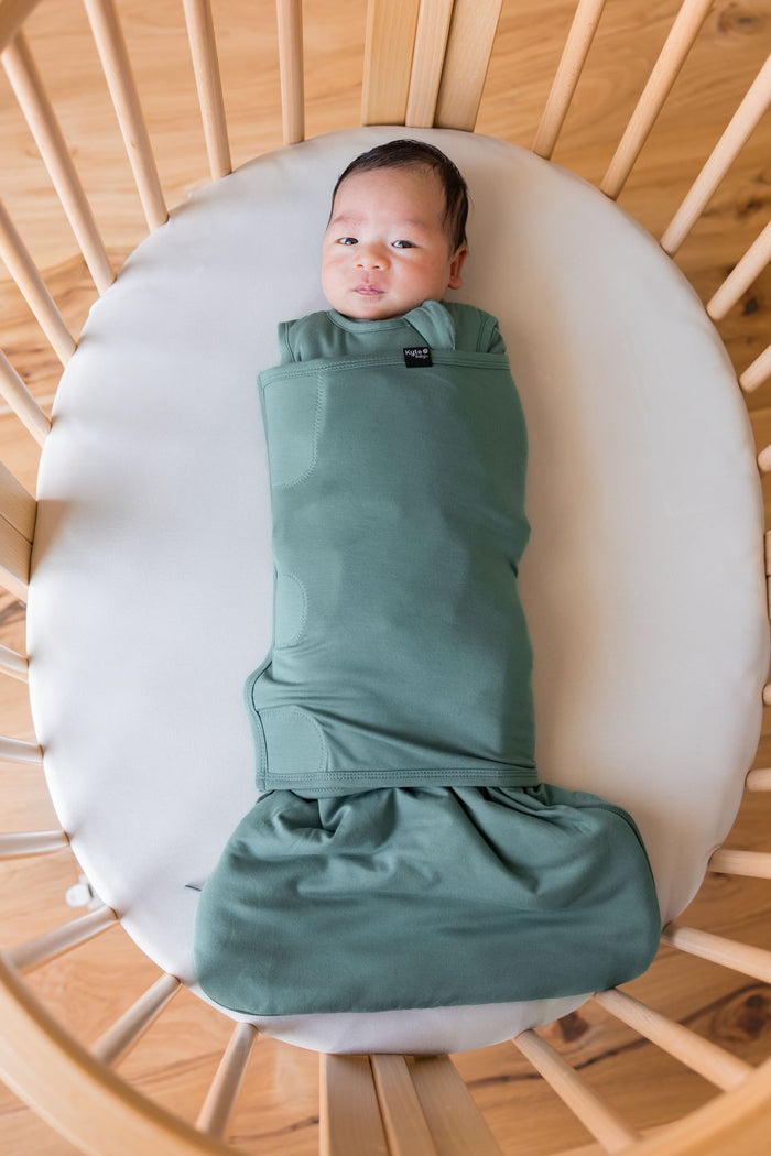 A baby swaddled in a dusty sage green swaddler, lying on a white mattress inside a wooden crib.