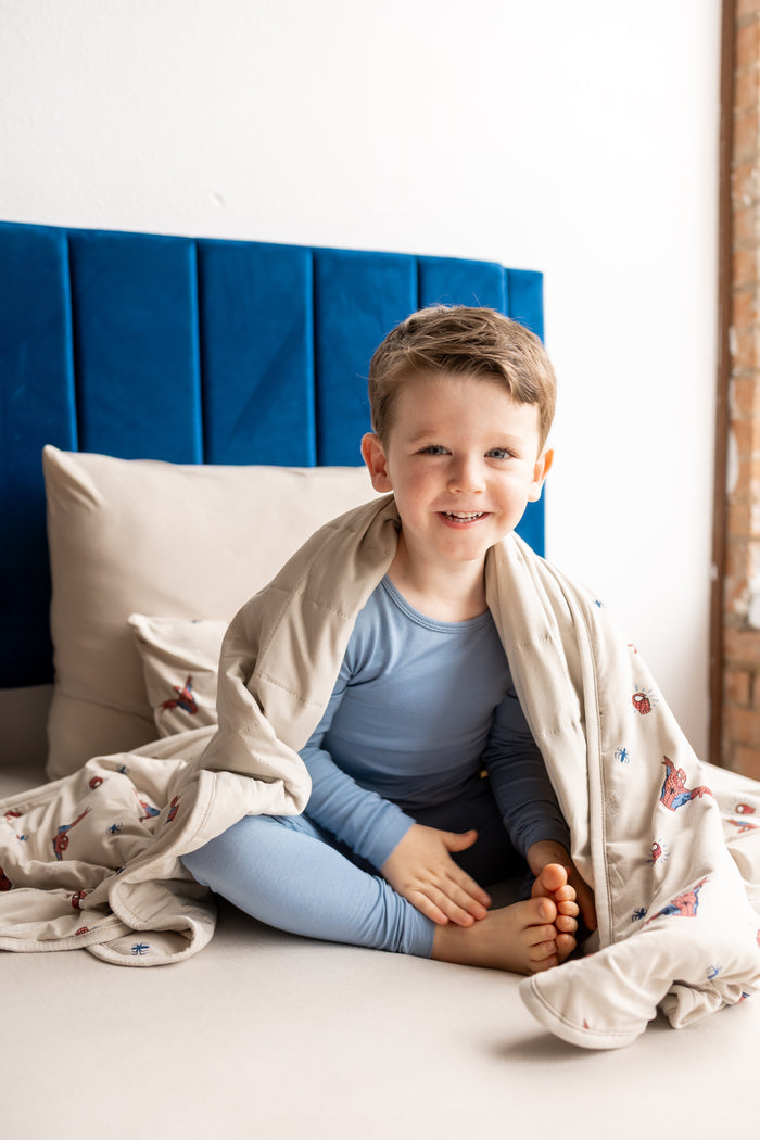 Boy sitting on bed wrapped in oat color Toddler Blanket with Spider-Man print wearing blue pajamas