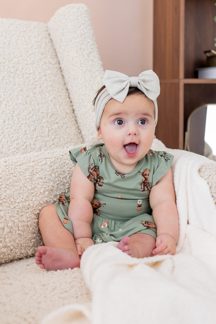 Baby smiling on a white sofa wearing Green Twirl Bodysuit Dress in Groot print and a beige knotted bow