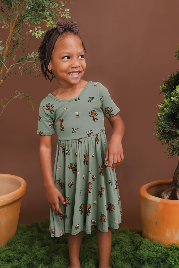 Girl smiling between plant pots wearing Green Twirl Dress in Groot print and brown bow